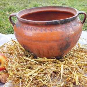 May include: A large, brown clay pot with two handles sits on a bed of straw. The pot has a decorative pattern around the rim and is slightly weathered.