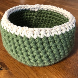 May include: A green crocheted bowl with a white crocheted trim. The bowl is made with a thick yarn and has a textured surface.