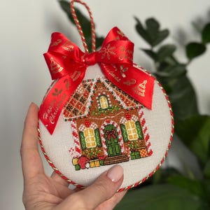 May include: A round Christmas ornament featuring a cross-stitched gingerbread house design. The ornament has a red and gold ribbon bow with "Happy Christmas" text. The edges are trimmed with gold and red cording.