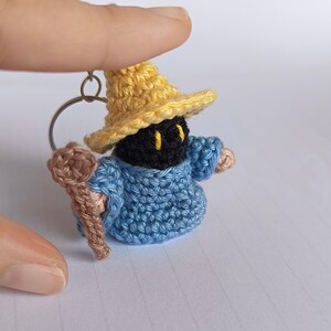 Könnte beinhalten: Eine gehäkelte Figur in Schwarz und Blau mit einem gelben Hut und einem braunen Stab. Die Figur ist an einem silbernen Schlüsselring befestigt.