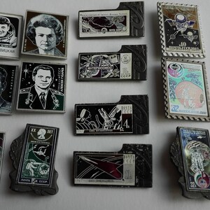 May include: A collection of rectangular metal pins featuring space-themed imagery, including portraits of cosmonauts, spacecraft, and Soviet-era stamps. The pins vary in shape, with some having decorative borders and others featuring Cyrillic text.