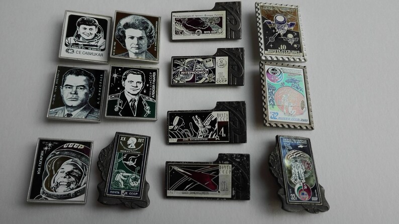 May include: A collection of rectangular metal pins featuring space-themed imagery, including portraits of cosmonauts, spacecraft, and Soviet-era stamps. The pins vary in shape, with some having decorative borders and others featuring Cyrillic text.