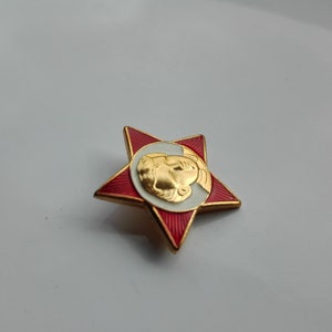K&ouml;nnte beinhalten: Eine rote sternf&ouml;rmige Anstecknadel mit goldfarbenem Rand und einem goldenen Profilportr&auml;t auf einem wei&szlig;en und goldenen kreisf&ouml;rmigen Hintergrund. Die Nadel hat einen goldenen Rand.