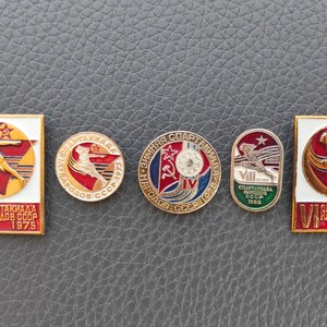 May include: Four vintage enamel badges featuring the Spartakiade, a Soviet sports festival. The badges are red, white, blue, and green with gold accents. The badges feature the text "Spartakiade" and "Narodov SSSR" (People of the USSR) and the years 1975 and 1983.