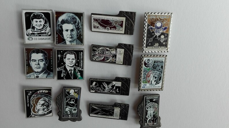 May include: A collection of twelve rectangular metal pins featuring Soviet space exploration themes, including portraits of cosmonauts, space station illustrations, and commemorative postage stamp designs with Cyrillic text and various graphic motifs.