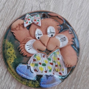 May include: A ceramic, round wall hanging featuring a cartoon rabbit. The rabbit has a brown body, a white and red bow, and a colorful shirt. The background is a mix of green and blue.