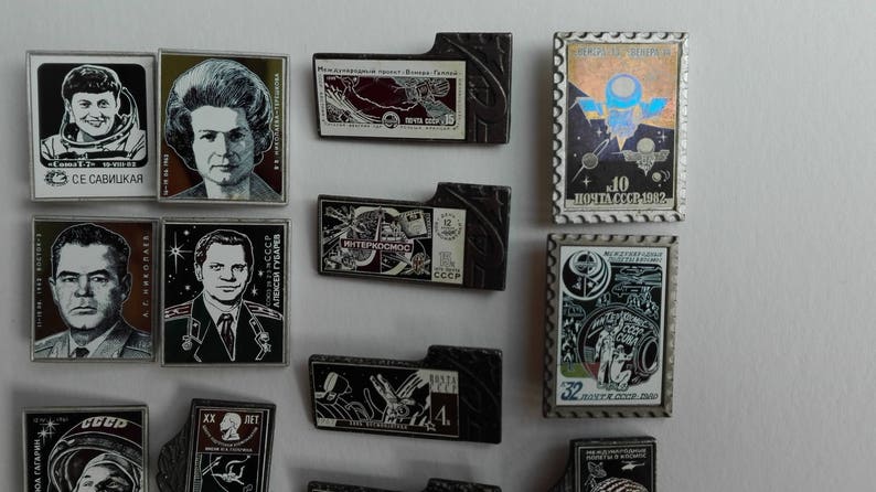 May include: A collection of rectangular metal pins featuring portraits of cosmonauts, space-themed illustrations, and Soviet postage stamp designs. The pins include Cyrillic text, black and white imagery, and colorful holographic accents on a white background.