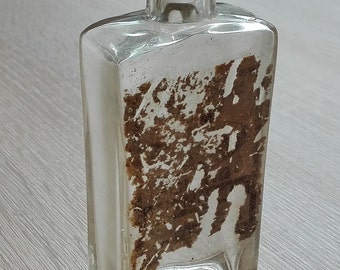 May include: A clear glass bottle with a square body and a round neck. The bottle has a brown label with faded text.