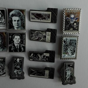 May include: A collection of twelve rectangular metal pins featuring space-themed imagery, including portraits of cosmonauts, spacecraft, and Cyrillic text. The pins vary in orientation and design, showcasing detailed black, white, and metallic graphics.