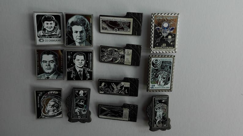 May include: A collection of twelve rectangular metal pins featuring space-themed imagery, including portraits of cosmonauts, spacecraft, and Cyrillic text. The pins vary in orientation and design, showcasing detailed black, white, and metallic graphics.