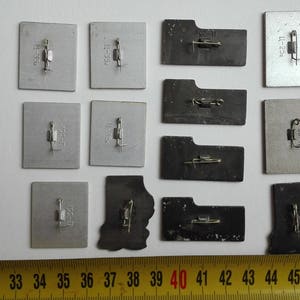 May include: A collection of twelve rectangular and irregular metal pins with pin-back fasteners attached to the reverse side. The pins are arranged in rows and columns, positioned above a yellow measuring tape showing measurements in centimeters.