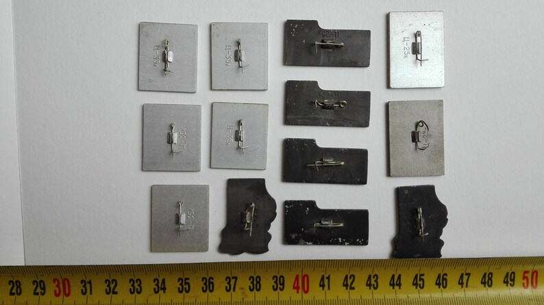 May include: A collection of twelve rectangular and irregular metal pins with pin-back fasteners attached to the reverse side. The pins are arranged in rows and columns, positioned above a yellow measuring tape showing measurements in centimeters.