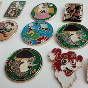 Könnte beinhalten: Eine Sammlung von 14 bunten Emaille-Anstecknadeln mit Comicfiguren und Tieren. Die Nadeln sind rund, oval und rechteckig. Einige der Nadeln haben Text, wie "GARDEN CITY" und "Жар-птица".
