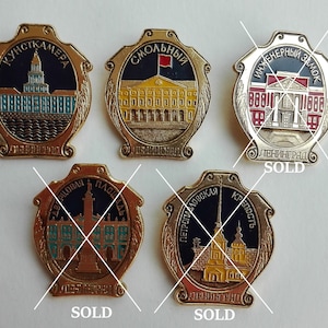 Puede incluir: Cinco pines de esmalte de color dorado que representan edificios famosos de San Petersburgo, Rusia. Los pines muestran la Kunstkamera, el Smolny, el Castillo de Ingeniería, el Almirantazgo y la Fortaleza de Pedro y Pablo. Cada pin tiene un fondo negro con letras doradas y un borde dorado.