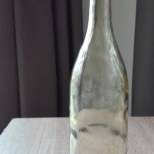 May include: A clear glass bottle with a slightly amber tint. The bottle has a rounded bottom and a tall, slender neck.