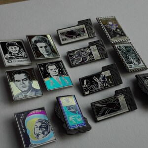 May include: A collection of rectangular metal pins featuring space-themed imagery, including portraits of cosmonauts, spacecraft, and celestial motifs. The pins display various black, white, and colored designs with Cyrillic text and dates.