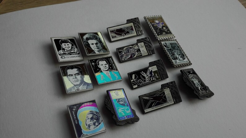 May include: A collection of rectangular metal pins featuring space-themed imagery, including portraits of cosmonauts, spacecraft, and celestial motifs. The pins display various black, white, and colored designs with Cyrillic text and dates.