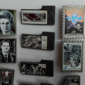 May include: A collection of rectangular metal pins featuring portraits of cosmonauts, space-themed illustrations, and Soviet postage stamp designs. The pins include Cyrillic text, black and white imagery, and colorful holographic accents on a white background.