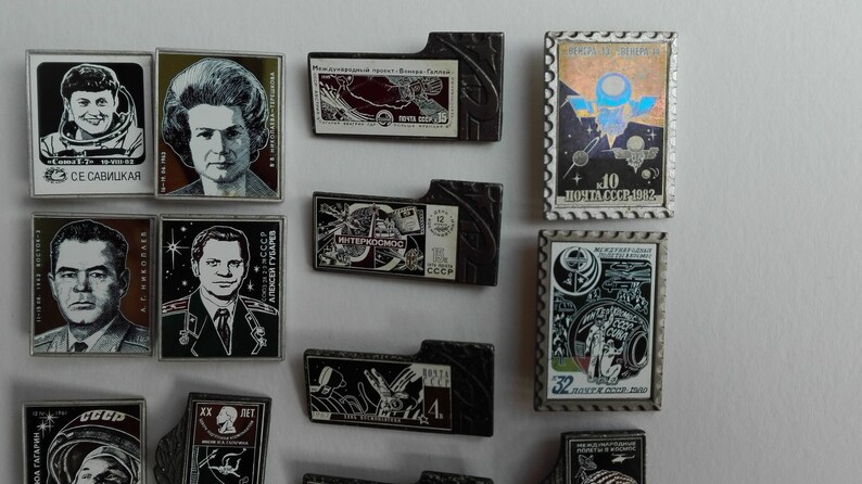 May include: A collection of rectangular metal pins featuring portraits of cosmonauts, space-themed illustrations, and Soviet postage stamp designs. The pins include Cyrillic text, black and white imagery, and colorful holographic accents on a white background.