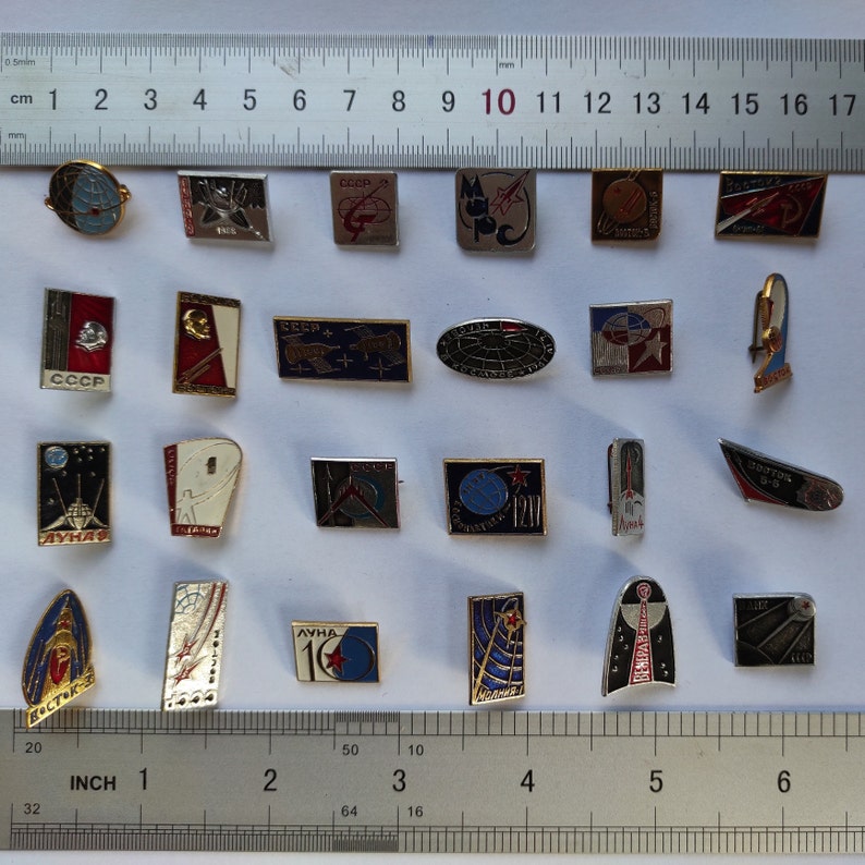 Peut inclure: Une collection de 20 &eacute;pingles en &eacute;mail vintage sur le th&egrave;me de l'exploration spatiale. Les &eacute;pingles sont dispos&eacute;es en rang&eacute;es sur une surface blanche avec une r&egrave;gle pour l'&eacute;chelle. Les &eacute;pingles pr&eacute;sentent des images de fus&eacute;es, de satellites, d'astronautes et de la Terre. Certaines &eacute;pingles comportent du texte, notamment "CCCP", "ЛУНА" et "ВОСТОК".