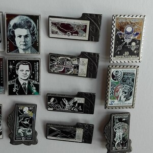 May include: A collection of twelve rectangular metal pins featuring Soviet space exploration themes, including portraits of cosmonauts, space station illustrations, and commemorative postage stamp designs with Cyrillic text and various graphic motifs.