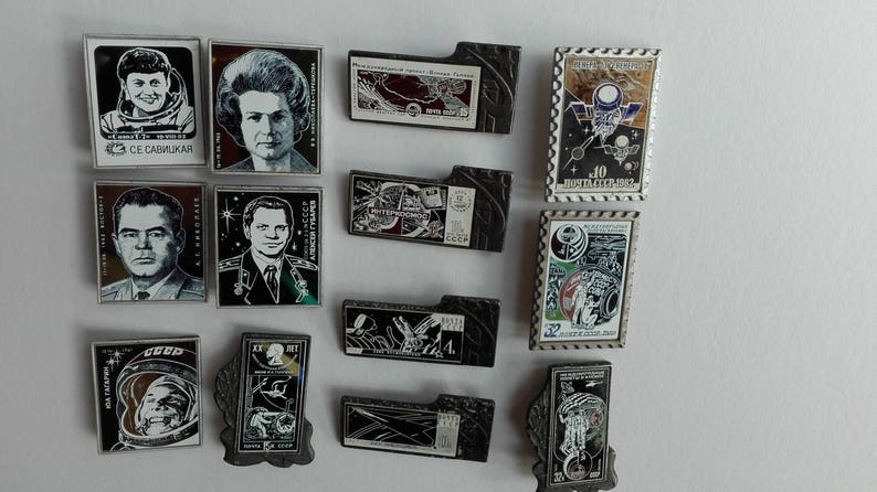 May include: A collection of twelve rectangular metal pins featuring Soviet space exploration themes, including portraits of cosmonauts, space station illustrations, and commemorative postage stamp designs with Cyrillic text and various graphic motifs.
