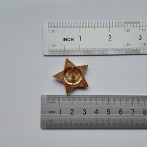 K&ouml;nnte beinhalten: Eine goldfarbene, sternf&ouml;rmige Anstecknadel mit einem Nadelverschluss. Die Anstecknadel hat einen Durchmesser von etwa 2,5 cm. Ein Lineal mit Zoll- und Zentimetermessungen befindet sich im Hintergrund.