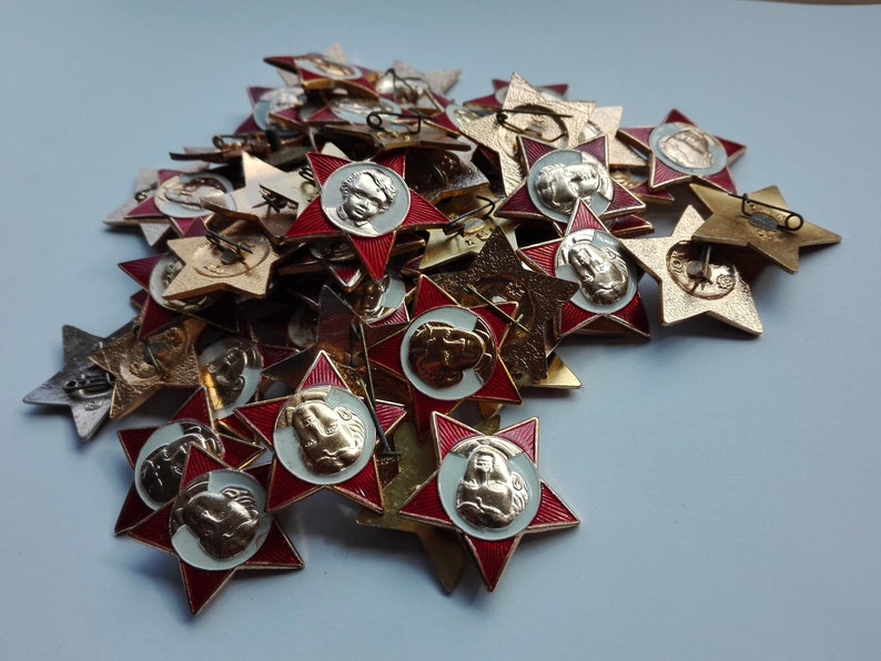 K&ouml;nnte beinhalten: Eine Sammlung von sternf&ouml;rmigen Vintage-Anstecknadeln. Die Anstecknadeln haben einen roten Emaille-Rand, ein zentrales Medaillon mit einem Reliefportr&auml;t und eine Anstecknadel. Die Anstecknadeln sind in verschiedenen Gold- und Silbert&ouml;nen gehalten.