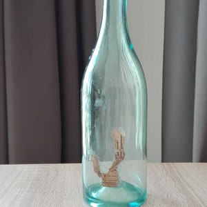May include: A clear glass bottle with a textured surface. The bottle is empty and has a cork stopper inside.