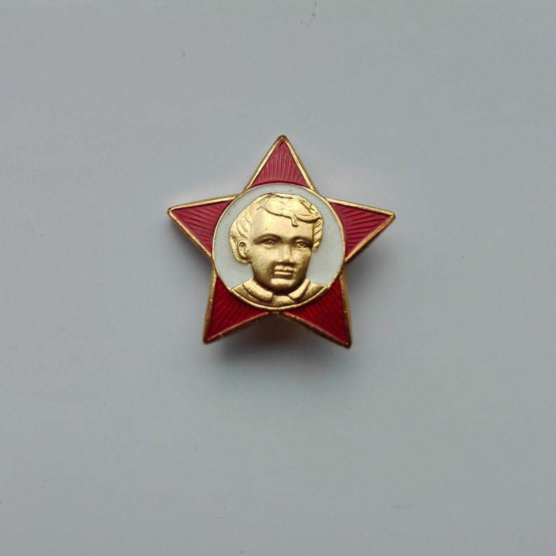 K&ouml;nnte beinhalten: Eine sternf&ouml;rmige Vintage-Anstecknadel mit roter Emaille und goldenen Verzierungen. Die Anstecknadel zeigt ein zentrales Medaillon mit einem goldfarbenen Portr&auml;t einer jungen Person vor wei&szlig;em Hintergrund. Der Stern hat eine rote Emaille-Oberfl&auml;che.