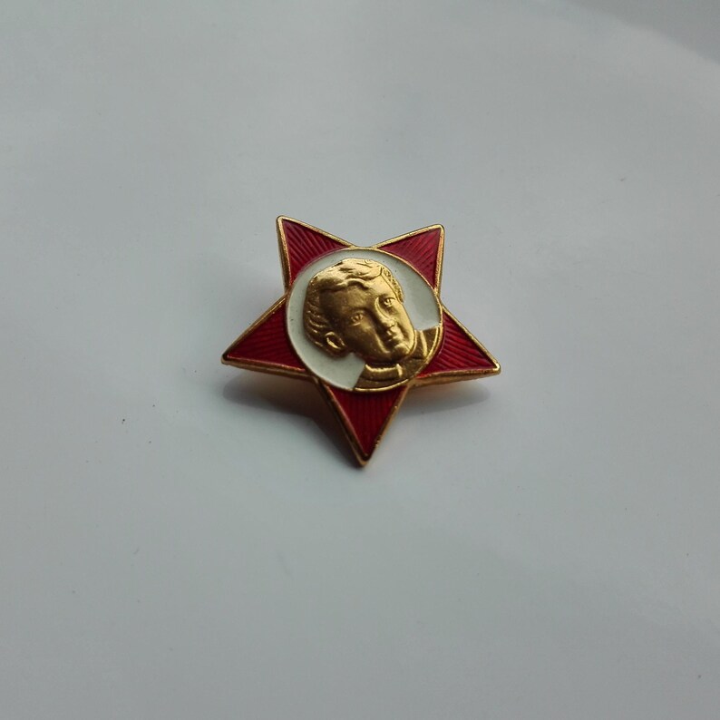 K&ouml;nnte beinhalten: Eine rote sternf&ouml;rmige Anstecknadel mit goldenem Rand und einem goldenen Portr&auml;t in der Mitte. Das Portr&auml;t ist von einem wei&szlig;en Kreis umgeben. Die Anstecknadel hat einen goldenen Rand und eine rote Emaille-Oberfl&auml;che.