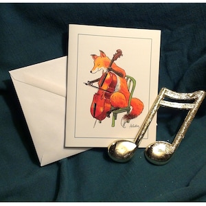 May include: A greeting card featuring a fox playing a cello. The fox is orange and white and is sitting on a chair. The cello is brown and has a gold string. The card is white with a gold border. The card is on a green surface with a gold musical note.