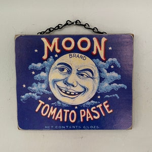 May include: A blue and white vintage sign with a cartoon moon face and the text "Moon Brand Tomato Paste" and "Net Contents 6 1/2 oz."