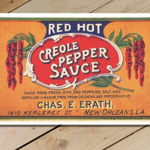 Könnte beinhalten: Vintage-Etikett für Red Hot Creole Pepper Sauce, hergestellt aus frischen, reifen roten Paprikaschoten, Salz und destilliertem Essig. Das Etikett zeigt ein rotes Oval mit dem Produktnamen und auf jeder Seite einen Bund roter Paprikaschoten. Das Etikett enthält auch den Firmennamen Chas. E. Erath und die Adresse 1410 Kerlerec St., New Orleans, LA.