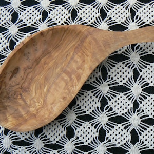 Op de afbeelding: Een houten serveerschep met een lange steel, gemaakt van olijfhout. De schep heeft een natuurlijk houtnerfpatroon en ligt op een wit gehaakt tafelkleed.