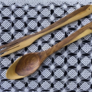 May include: Two wooden kitchen utensils, a spoon and a fork, made from olive wood, on a white crocheted tablecloth.