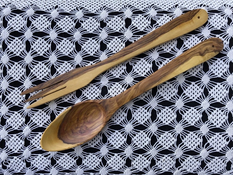 May include: Two wooden kitchen utensils, a spoon and a fork, made from olive wood, on a white crocheted tablecloth.
