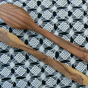 May include: A set of two wooden kitchen utensils, a spoon and a fork, made from olive wood. The utensils are laid out on a white crocheted tablecloth.