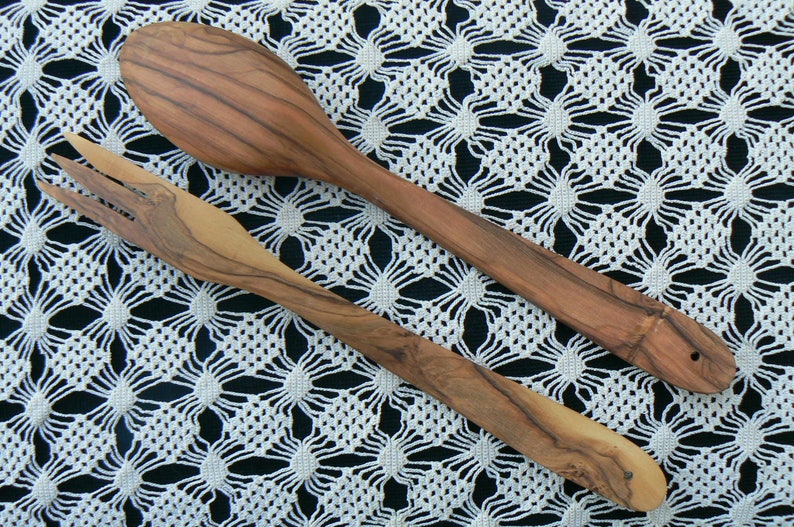 May include: A set of two wooden kitchen utensils, a spoon and a fork, made from olive wood. The utensils are laid out on a white crocheted tablecloth.