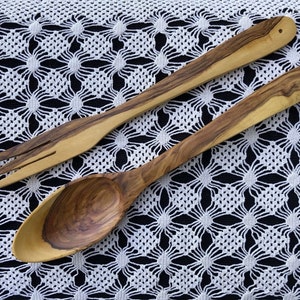 May include: A wooden spoon and fork set made from olive wood. The utensils are laid out on a white crocheted tablecloth.