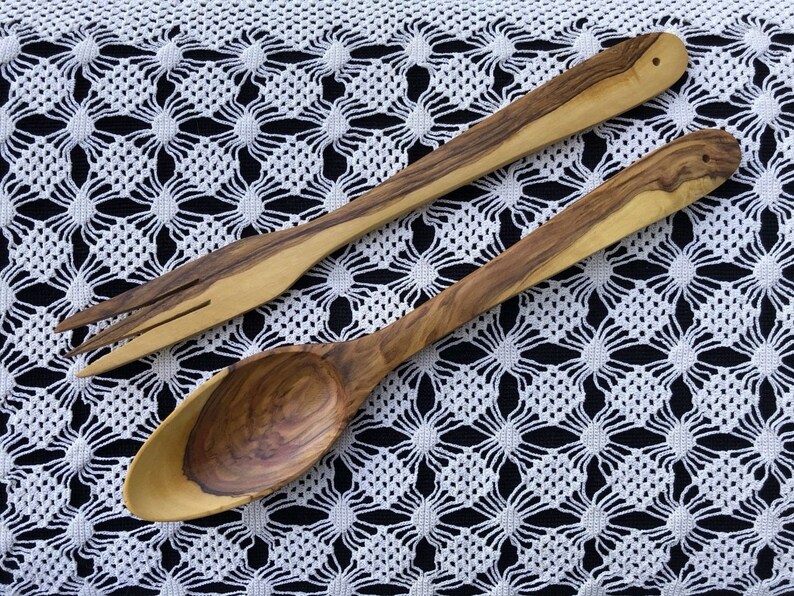 May include: A wooden spoon and fork set made from olive wood. The utensils are laid out on a white crocheted tablecloth.