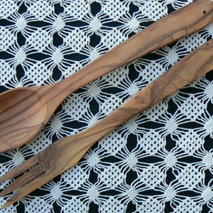 May include: Two wooden kitchen utensils, a spoon and a fork, are laid out on a white crocheted tablecloth. The utensils are made of light brown wood with a natural grain pattern.