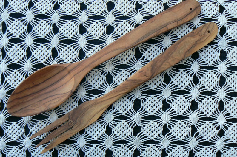 May include: Two wooden kitchen utensils, a spoon and a fork, are laid out on a white crocheted tablecloth. The utensils are made of light brown wood with a natural grain pattern.