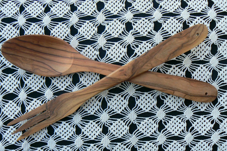 May include: A wooden salad serving set with a spoon and fork. The utensils are crossed and laid on a white crocheted tablecloth.