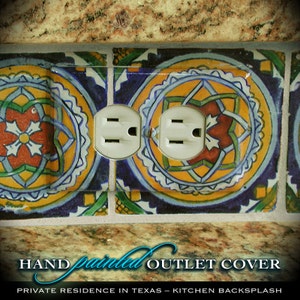May include: Hand-painted outlet cover with a decorative design. The cover features a vibrant pattern of yellow, blue, and red floral motifs. The text "Hand Painted Outlet Cover" is displayed at the bottom. The outlet cover is installed in a kitchen backsplash.