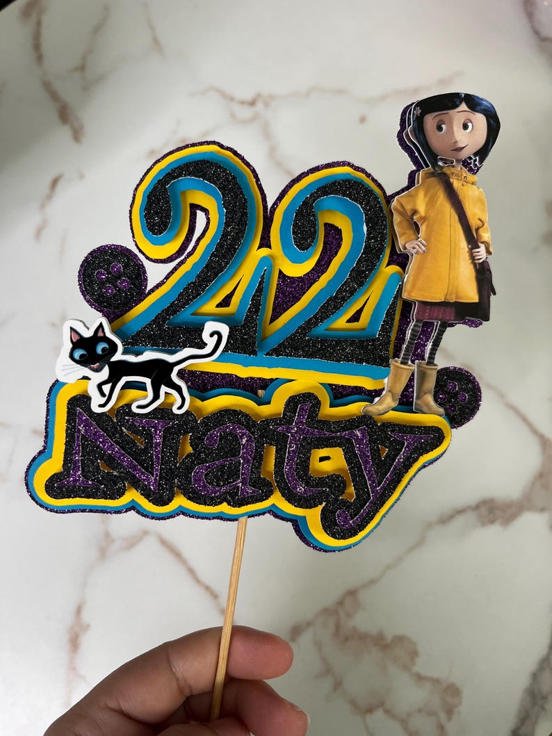 May include: A cake topper featuring a yellow-clad character with black hair and a black cat. The number 22 is in the background with a purple, blue, and yellow glitter design. The name "Naty" is also included in the design.