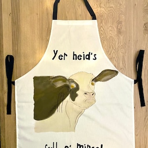 以下が含まれることがあります： 黒い牛のイラストと「Yer heid's full o' mince!」というテキストが書かれた白いキッチンエプロン