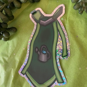 Pode incluir: Um autocolante com um design de avental verde escuro com uma contorno verde claro e uma borda de glitter prateado. O avental tem um pequeno gráfico de regador. O autocolante está sobre um fundo verde com vegetação artificial.