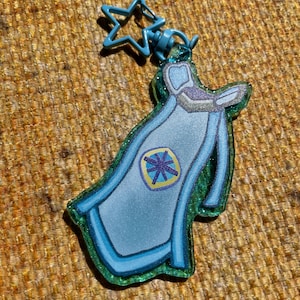 Pode incluir: Um chaveiro azul e verde com um design de capa e um fecho em forma de estrela. A capa tem um emblema central com um design roxo e amarelo. O chaveiro é feito de um material brilhante.