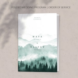 Op de afbeelding: Een gevouwen huwelijksvoltrekkingsprogramma met een aquarelillustratie van een mistig bos en bergen. De tekst luidt "Welkom op de bruiloft van Maya Harris & Oliver Bailey 05-07-25".